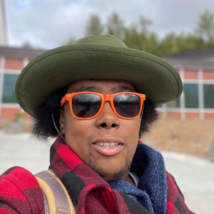 A selfie of a Black person standing in front of a one-story building, low trees, and a partly cloudy sky in a wide-brimmed, army-green hat, orange sunglasses, and red flannel coat.