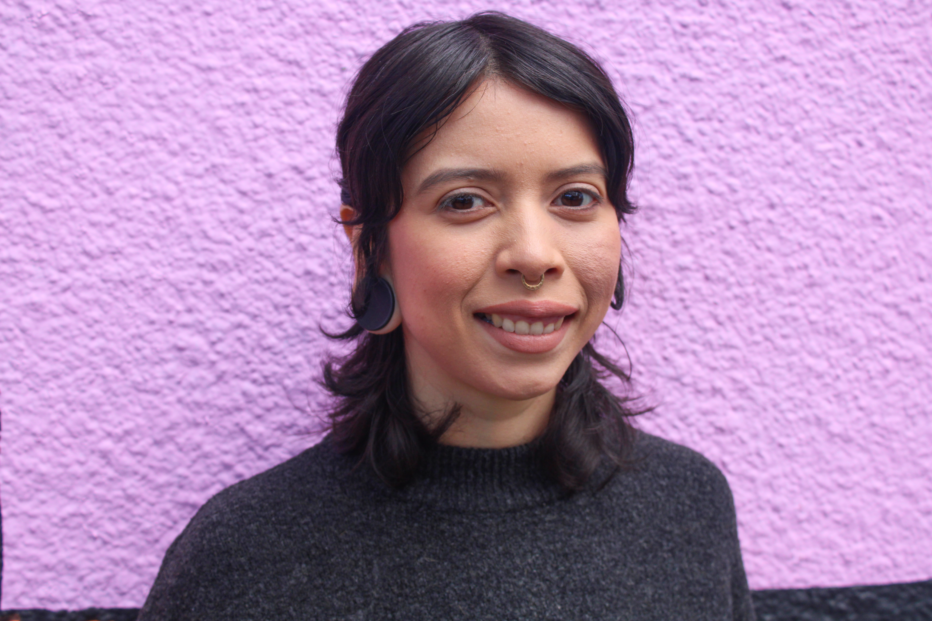 A Latine person with shoulder-length brown hair that curls out at the ends, and with large black plugs in their earlobes and a septum ring stands in front of a textured light pink-purple wall in a dark gray sweater.