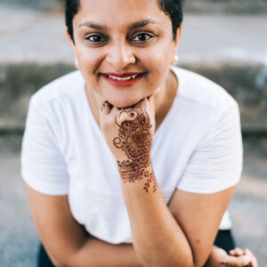 A South Asian woman with short, dark hair sits smiling in a white t-shirt and black pants, with her mendhi-decorated hand under her chin.